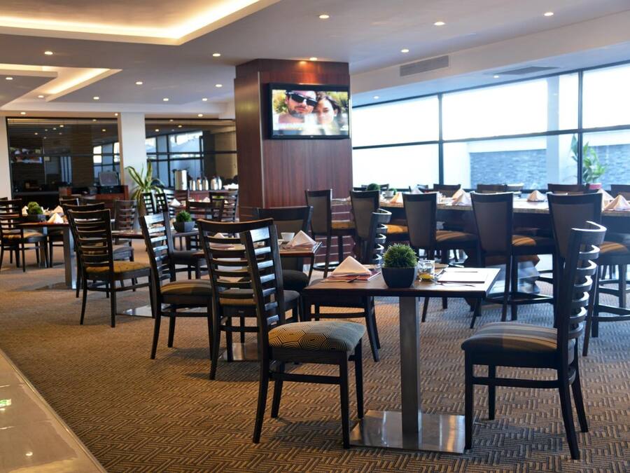 Un comedor de hotel con varias mesas y sillas, un televisor instalado en un pilar, y ventanas amplias que ofrecen vistas al exterior.