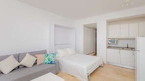A bedroom with a bed, a grey sofa with patterned cushions, a white bedside table, and a kitchenette with a microwave and refrigerator.