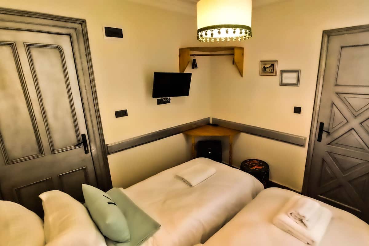 Una habitación pequeña y funcional con dos camas, un escritorio, una silla, una lámpara y un televisor.