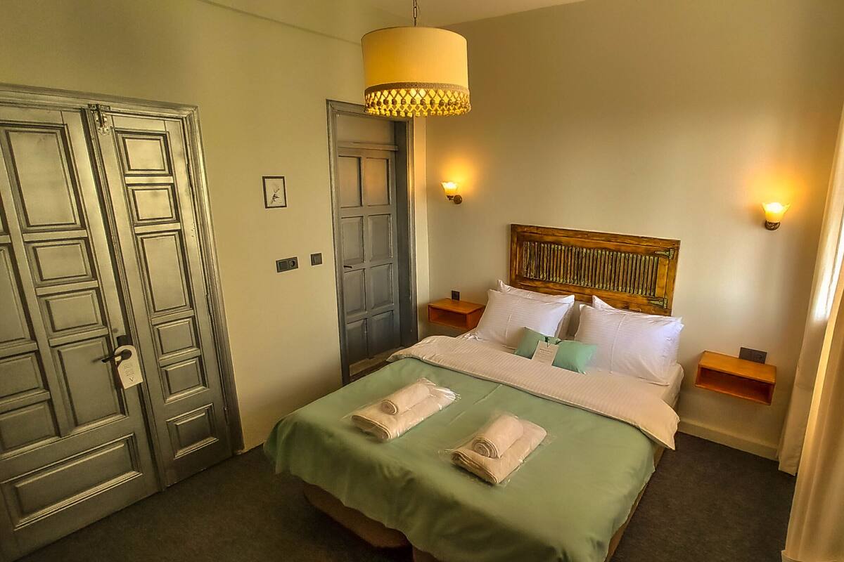 Habitación de hotel con cama, dos mesitas de noche, cabecera de madera, lámpara colgante y puerta con cerradura.