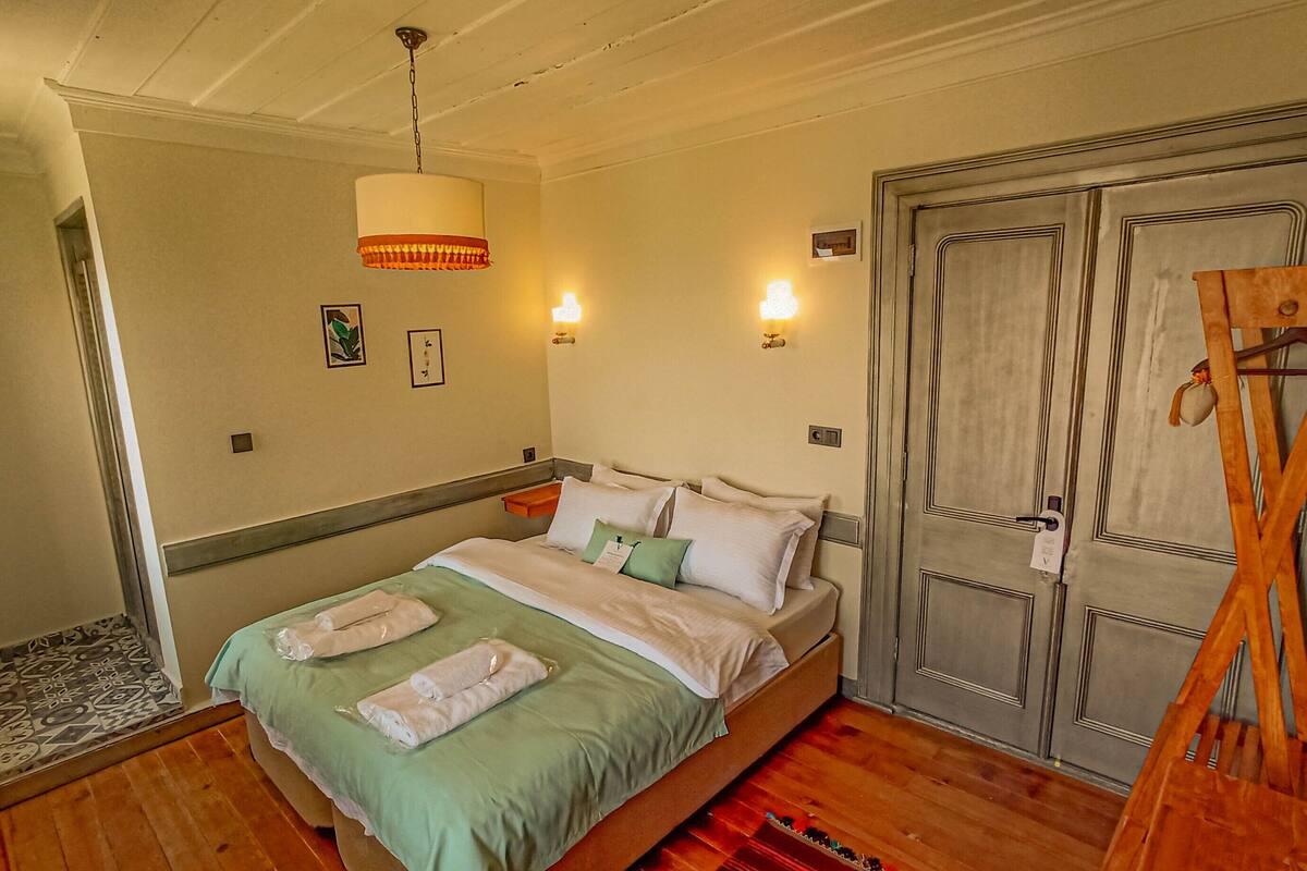 Una cama bien hecha con almohadas y una colcha, suelo de madera, lámpara colgante y un armario de madera.