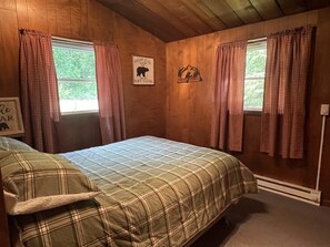 Quarto de madeira com uma cama, duas janelas com cortinas xadrez, decoração de urso e montanha nas paredes e uma moldura com silhueta de urso.