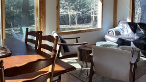 A cozy cabin interior with a wooden dining table, chairs, a sofa, and large windows offering views of a forested area. - Premier Cottage Getaway-Acorn Grove, Pentwater, MI (Pentwater)