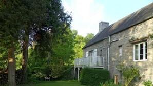 A stone house with a wooden deck, surrounded by lush greenery and a well-maintained lawn. - Gite de la petite suisse "BAMBOU" (Clécy)