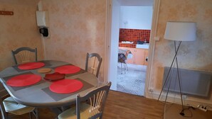 A dining area with a table set for four, chairs, a lamp, and a view into a kitchen with red tiles. - Gite de la petite suisse "BAMBOU" (Clécy)