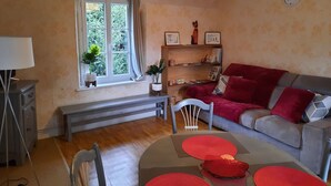 A cozy living room with a sofa, a coffee table, a lamp, a bookshelf, and a window with curtains. - Gite de la petite suisse "BAMBOU" (Clécy)