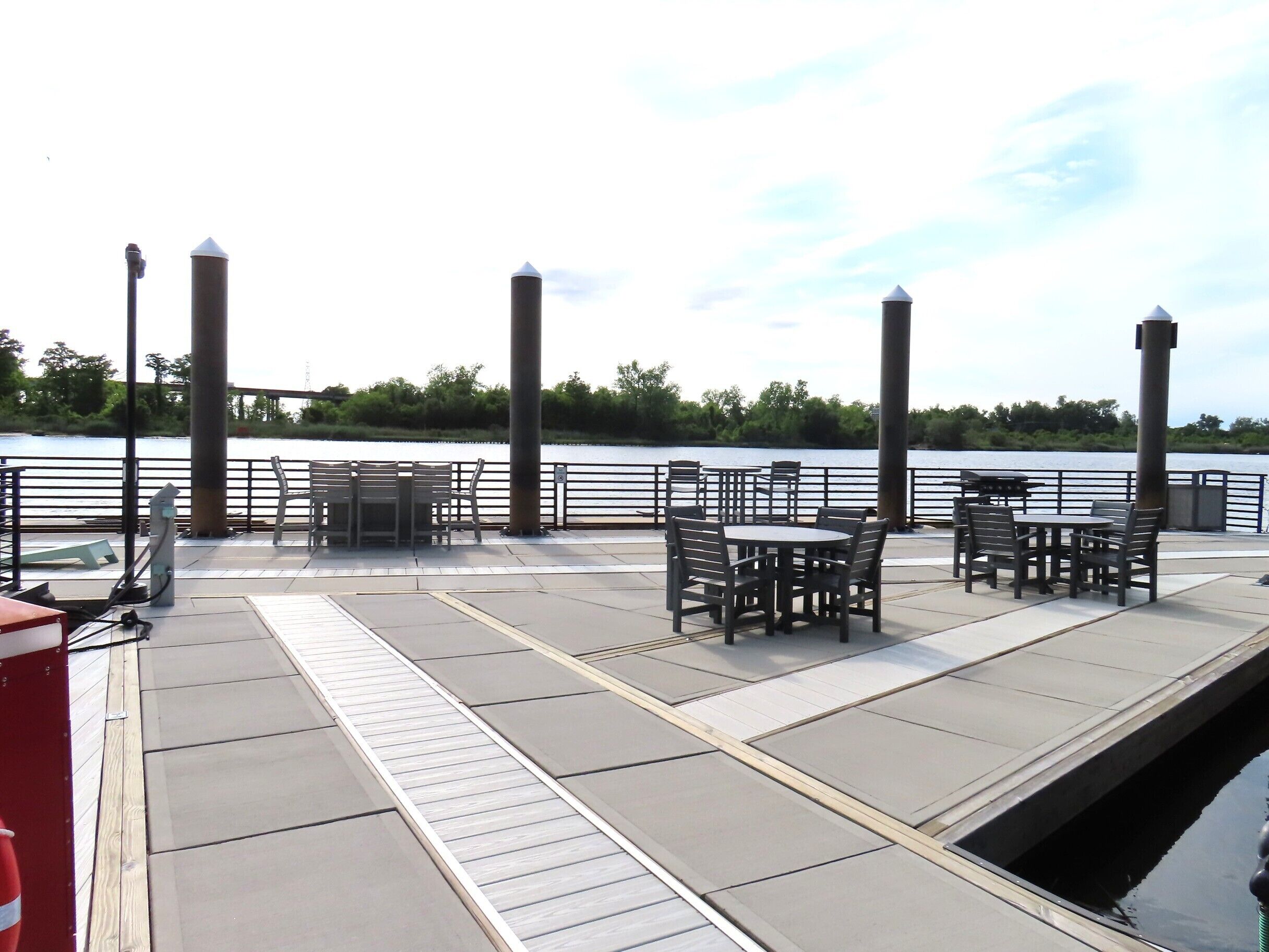 A riverside patio with tables and chairs, a red fire extinguisher, and a bridge in the background.