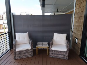 En balkon med to fletstole og et lille bord, en mørk væg og udsigt til bygninger.