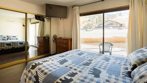 Un dormitorio con una cama grande, un aparador, un televisor montado en la pared y un ventanal corredero que da a un balcón con vistas a un edificio y coches aparcados.