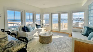 Un vaste salon doté d’une grande fenêtre donnant sur une plage, d’un canapé blanc agrémenté de coussins à motifs, d’une table basse ronde et d’un coin repas avec une table en marbre.