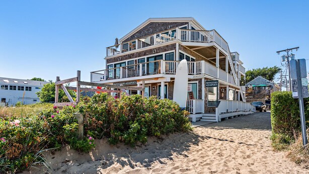 Un edificio frente a la playa con varios pisos y balcones, una playa arenosa al frente, y un letrero que dice "Provincetown Aquasports".
