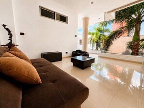 A modern living room with a sofa, ottoman, and coffee table. There is a large window with a view of palm trees and a building.