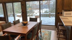 A dining area with a wooden table and chairs, a bowl of fruit, and a view of a snowy outdoor deck through large windows.