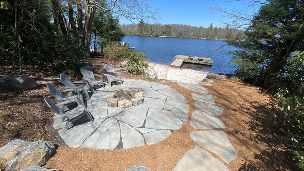 A stone patio with Adirondack chairs and a fire pit, overlooking a lake.