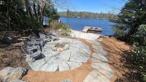 Un patio de piedra con sillas Adirondack y un hogar para fuego, con vista a un lago.