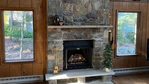 A stone fireplace with a mantel, flanked by two windows with a view of trees.