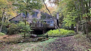 A two-story cabin surrounded by dense forest, with a covered deck and a chimney.