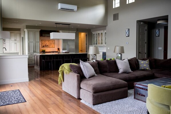A modern living room with a brown sectional sofa, a small yellow chair, and a white rug. A kitchen with white cabinets and a central island is visible in the background.