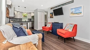 Uma sala de estar moderna com um sofá, duas poltronas vermelhas, uma TV fixada na parede e uma cozinha ao fundo.