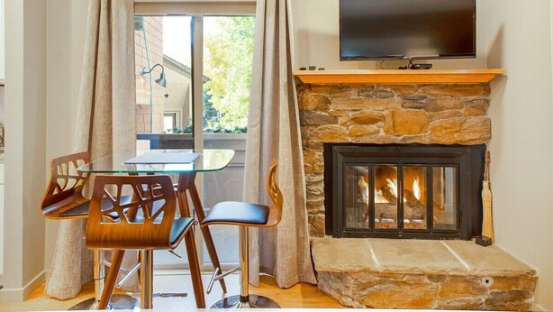 A cozy living room with a stone fireplace, a flat-screen TV mounted above it, and a dining area with a glass table and wooden chairs. - Elkhorn Condo Walk to Dollar! (Sun Valley)