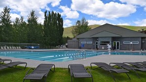 A swimming pool with lounge chairs, a building with a sloped roof, and a grassy area with trees in the background. - Elkhorn Condo Walk to Dollar! (Sun Valley)
