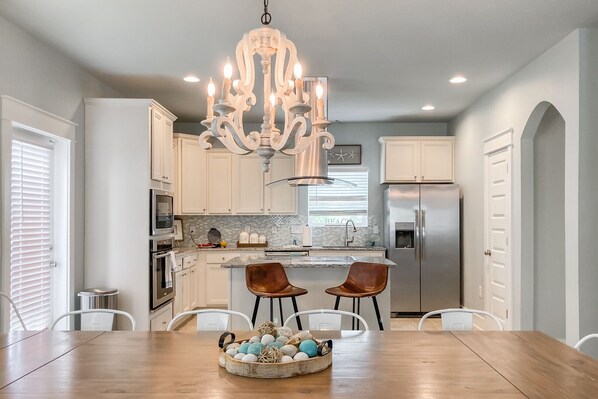A modern kitchen with white cabinets, a stainless steel refrigerator, a chandelier, and a dining area with a wooden table and two chairs.