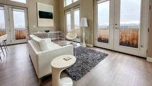 Uma sala de estar espaçosa com um sofá branco, uma mesa redonda pequena e uma janela grande com vista para uma cerca de madeira.