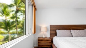 Un dormitorio con cabecera de madera, una mesita de noche con lámpara y vistas a palmeras a través de una ventana.