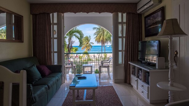 A living room with a view of the beach, a sofa, a coffee table, a TV, and a lamp. - Sandcastles Bed & Breakfast Hotel (Ocho Rios)