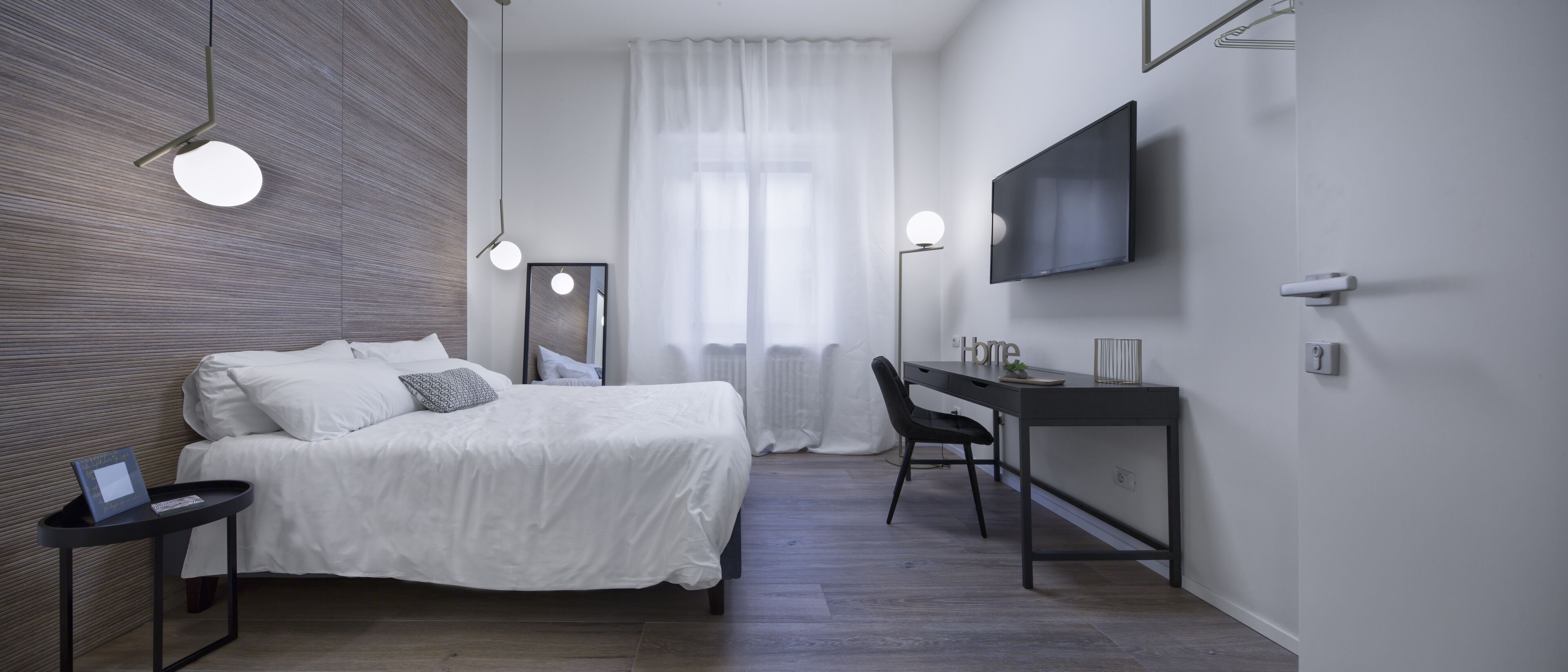 Quarto de hotel moderno com cama, mesa de cabeceira, cadeira, escrivaninha, televisão e janela com cortinas.