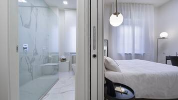 Habitación de hotel moderna con cabina de ducha de vidrio, cama con ropa blanca, escritorio con televisor de pantalla plana y vista a la ciudad a través de cortinas traslúcidas.