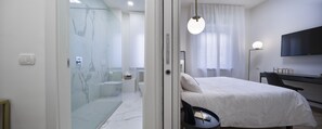 A modern hotel room with a glass shower enclosure, a bed with white bedding, a desk with a flat-screen TV, and a view of the city through sheer curtains.