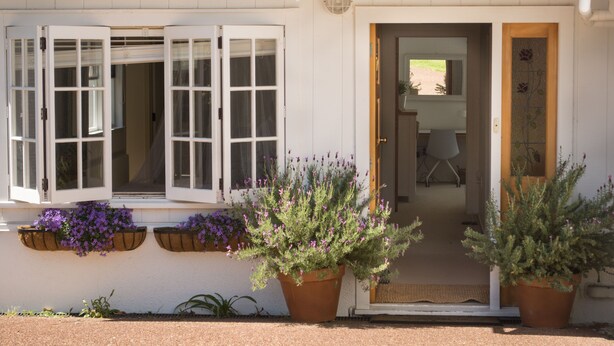A white building with a door and windows, potted plants, and a gravel driveway. - Oneroa Escape - Bell's Cottage (Waiheke Island)