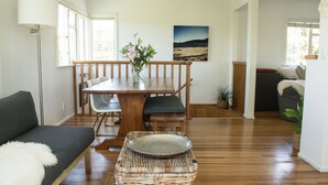 A modern living room with a wooden dining table, a sofa, and a wicker ottoman. - Oneroa Escape - Bell's Cottage (Waiheke Island)