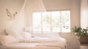 A bedroom with a bed, a bedside table, a lamp, a window with blinds, and a wall decoration. - Oneroa Escape - Bell's Cottage (Waiheke Island)