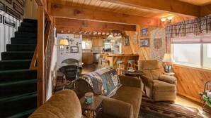 Un intérieur de chalet chaleureux avec un escalier, un salon doté d’un canapé et de fauteuils, un coin cuisine et un espace repas.