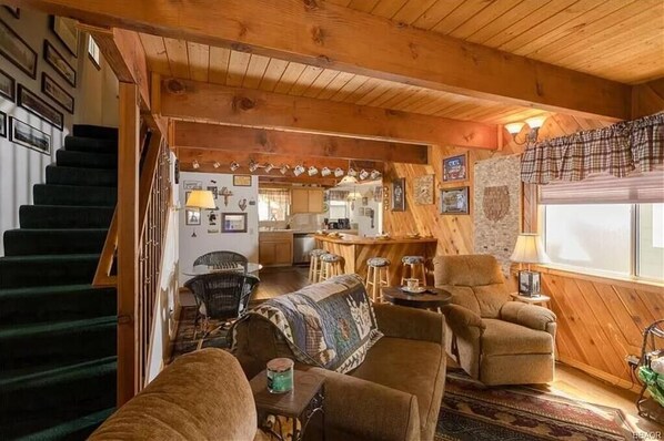 A cozy cabin interior with a staircase, a living room with a sofa and armchairs, a kitchen area, and a dining space. - Rustic Mountain Retreat. Trail access, fenced yard (Big Bear Lake)