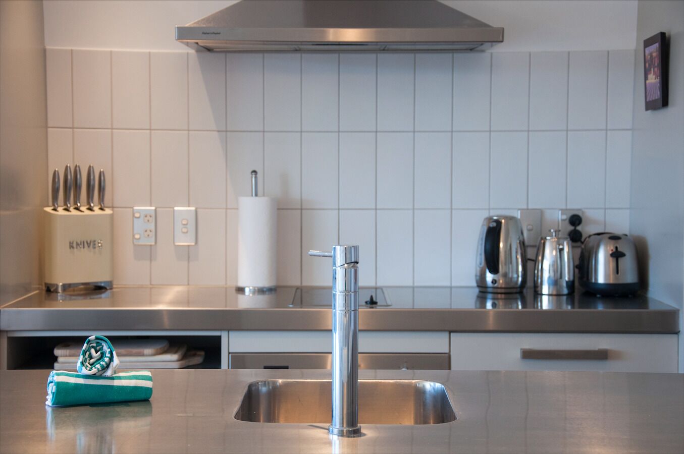 A modern kitchen with a stainless steel sink, a knife block, a paper towel dispenser, and a kettle.