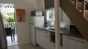 A compact kitchen with white cabinets, a built-in oven, a microwave, and a sink. There's a window above the sink and a door leading to a balcony with a table and chairs. - Kaymargo, Creole bungalow (Saint-Esprit)