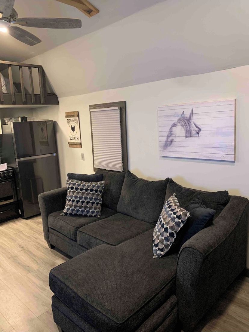 A living room with a dark grey sectional sofa, two patterned throw pillows, a wall-mounted artwork of a horse, a kitchen area with a refrigerator, and a ceiling fan.
