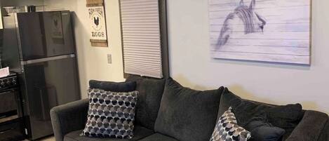 A living room with a dark grey sectional sofa, two patterned throw pillows, a wall-mounted artwork of a horse, a kitchen area with a refrigerator, and a ceiling fan.