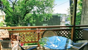A wooden deck with a glass table and chairs, a wooden bench with cushions, and a view of a green house in the background. - Panoramic view, large deck, ground floor apartment, walk to subway and beach (Boston)