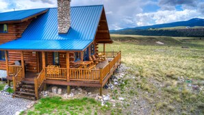 A log cabin with a blue metal roof, a stone chimney, and a wooden deck with outdoor furniture. - True Montana Log Cabin, Yellowstone Park, Ennis, Madison River Fishing,  4+2.5 (Cameron)