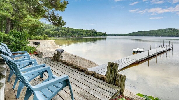 A lakeside deck with blue chairs, a wooden dock, and a calm lake. - Private Beach! Kayak, Swim, Fish! Hot Tub, Sauna & Boat Dock. Year-Round Fun! (Harrison)