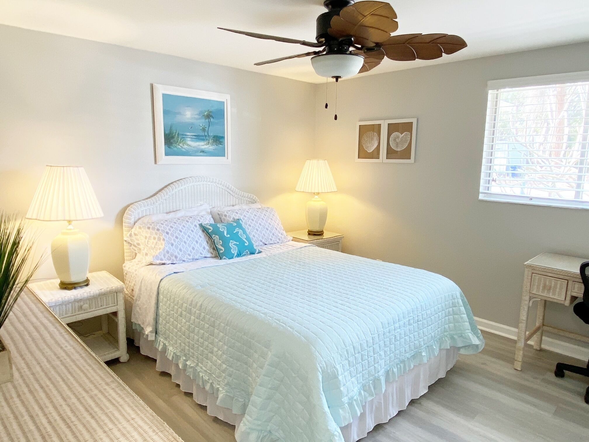 A bedroom with a bed, two bedside tables with lamps, a ceiling fan, a window with blinds, and wall art.