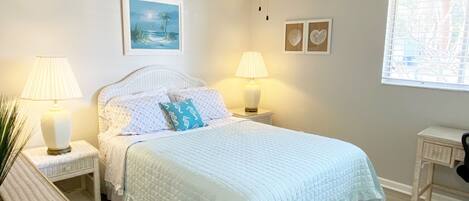 A bedroom with a bed, two bedside tables with lamps, a ceiling fan, a window with blinds, and wall art.