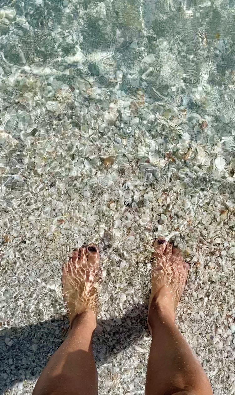 En persons fötter står på en stenstrand med klart vatten.