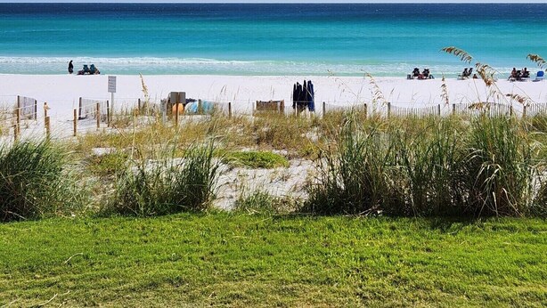 Une plage de sable blanc, avec une eau turquoise et limpide, où quelques personnes se détendent. On aperçoit une clôture en bois au premier plan.
