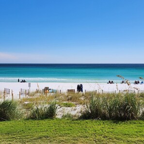 A beach with white sand, clear turquoise water, and a few people relaxing. There is a wooden fence in the foreground.