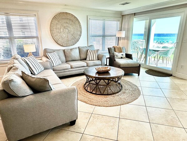 A spacious living room with a grey sectional sofa, a round coffee table, and a view of the beach through large windows.